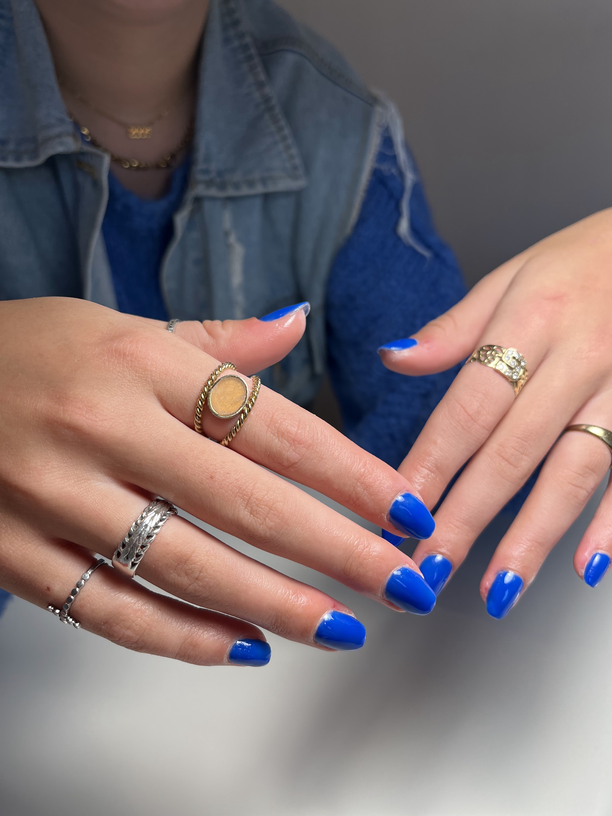 Twee handen met kobaltblauwe gelnagels en zilveren ringen, over een spijkerjasje